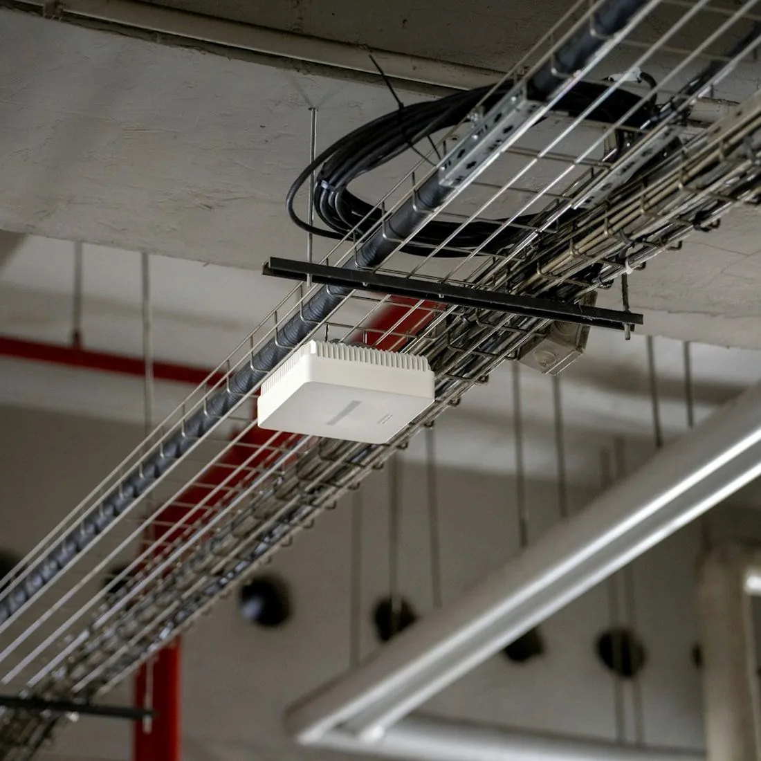 White Wi-Fi access point hanging from a wire mesh cable tray under a building ceiling